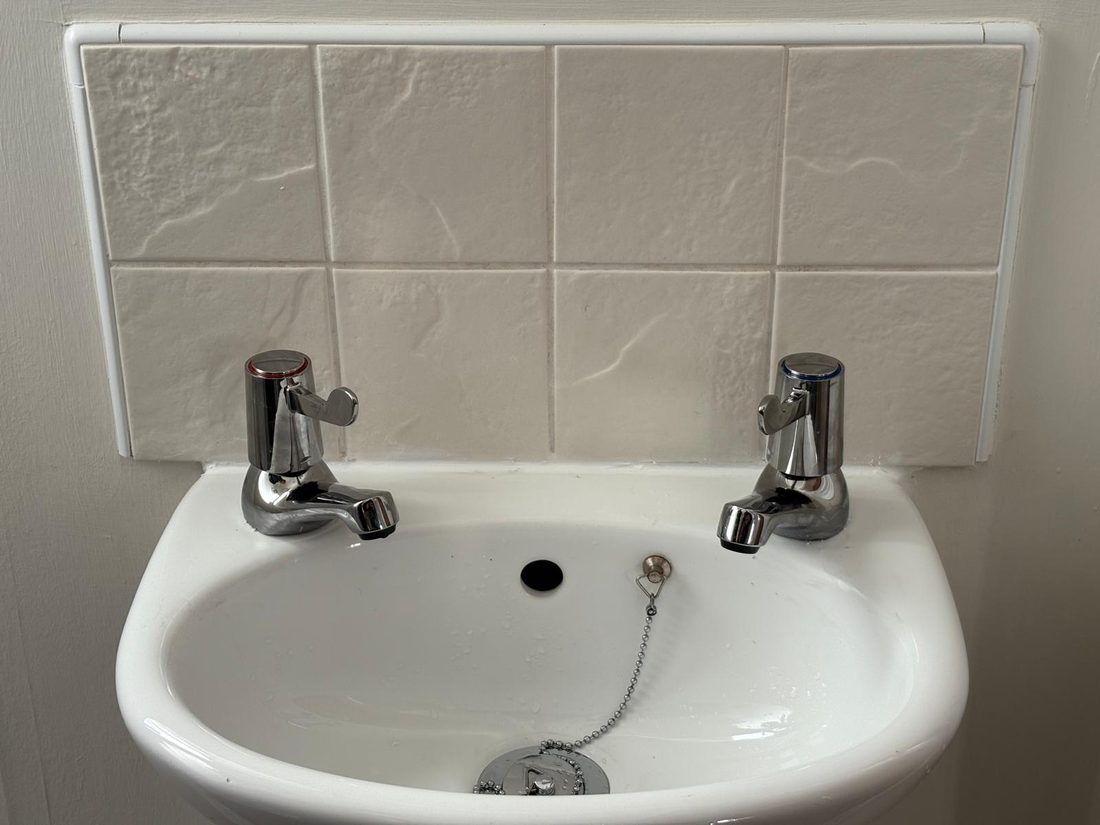 A small white basin with two chrome taps and a tiled splashback
